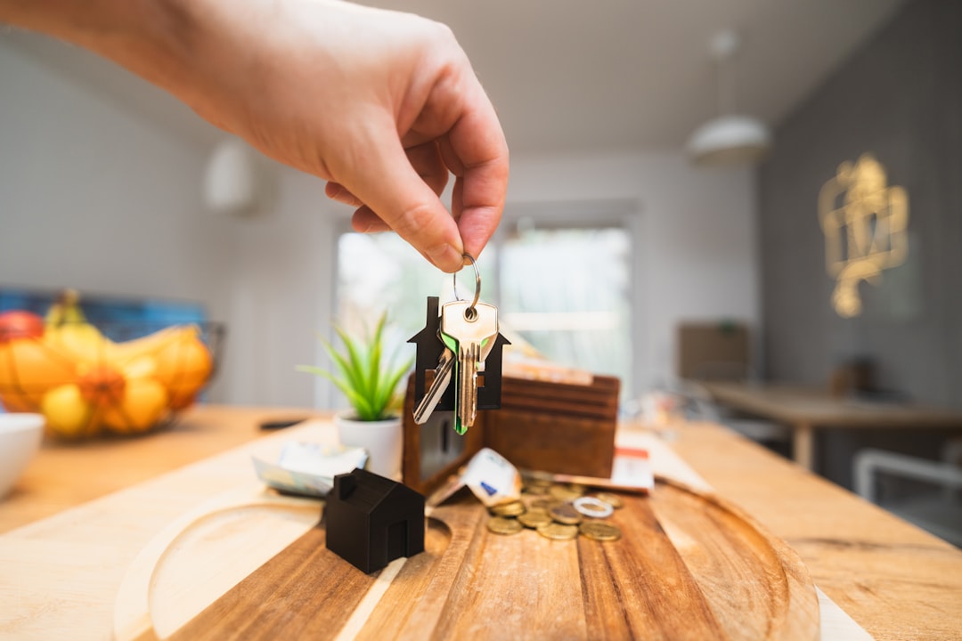 A person's hand carefully places a coin into a white, house-shaped piggy bank on a clean background, representing saving money on a home sale.