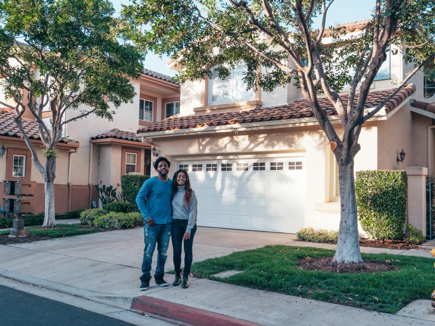 A smiling, diverse couple stands proudly in front of their beautiful, modern suburban house on a sunny day, symbolizing a successful home sale.